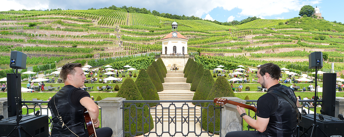 Zwei Musiker mit Gitarren spielen auf dem Schlossbalkon mit Blick auf das Belvedere von Schloss Wackerbarth und die umliegenden Weinberge.