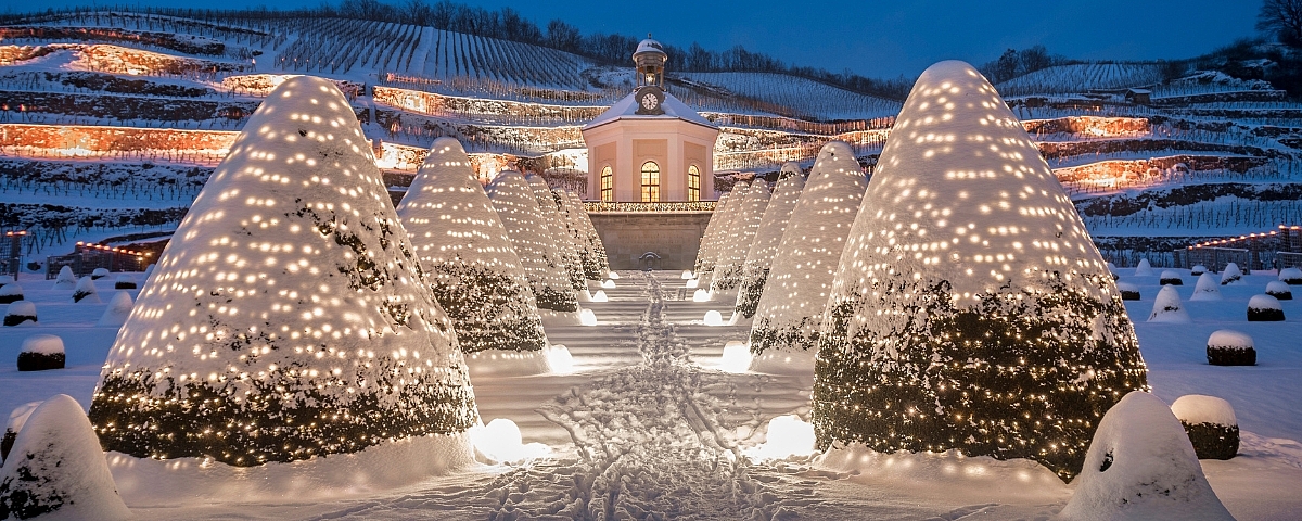 Festlich beleuchtete Gartenanlage von Schloss Wackerbarth im Schnee mit Blick auf das Belvedere, umgeben von winterlich beleuchteten Weinbergen.