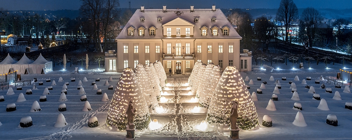 Winterwanderung auf Schloss Wackerbarth Schloss Wackerbarth im Schnee bei Nacht, festlich beleuchtet mit Lichterketten und winterlicher Gartenanlage.