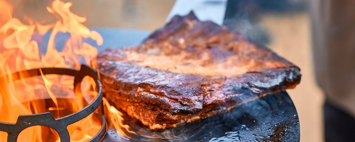 Wackerbarths Grillgenuss Großes Stück Fleisch wird am offenen Feuer gegrillt, mit knuspriger Kruste und sichtbaren Flammen.