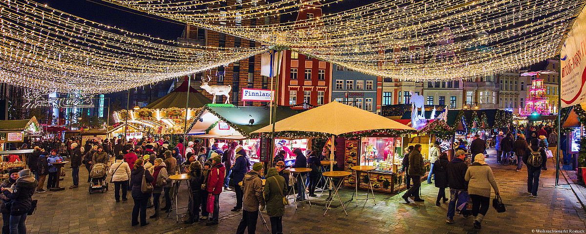 Zu sehen ist der hell erleuchtete Weihnachtsmarkt Rostock mit ganz vielen kleinen Ständen. Auch Schloss Wackerbarth ist vertreten. 