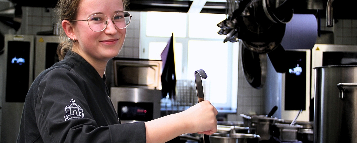 Azubi Koch / Köchin  Eine Auszubildende oder junge Köchin bei Schloss Wackerbarth steht in einer professionell ausgestatteten Küche und arbeitet mit einem Küchenwerkzeug. Ihr freundlicher Blick und die hochwertige Umgebung zeigen den praxisnahen, modernen Ausbildungsalltag im Bereich Gastronomie.