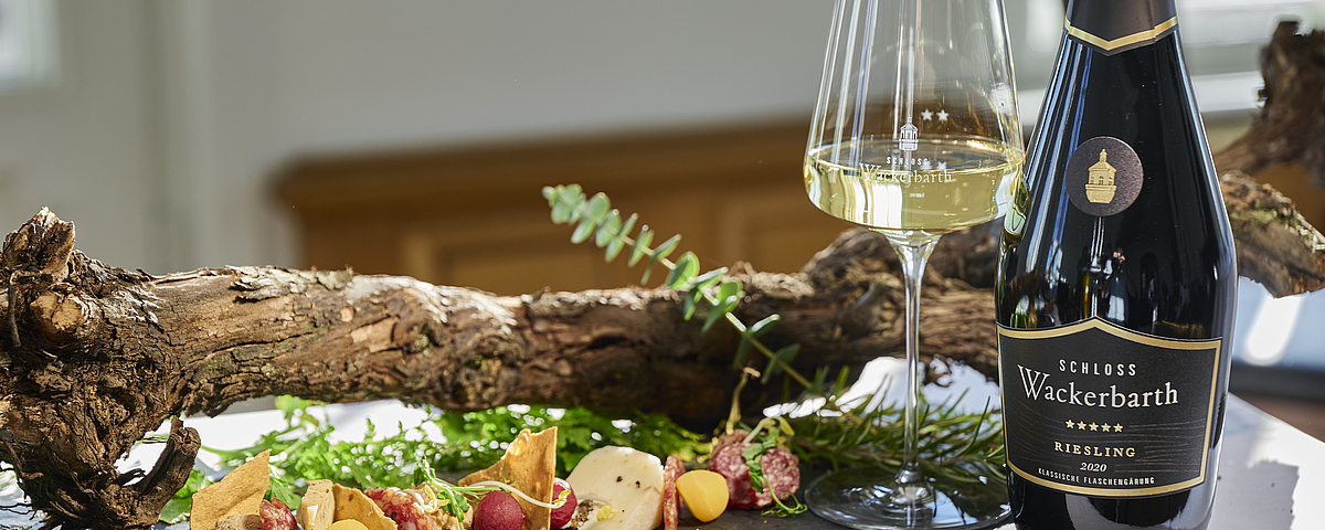 Eine Flasche Schloss Wackerbarth Riesling 2020 steht neben einem gefüllten Weinglas auf einem Tisch. Davor liegt eine Schieferplatte mit Käse, Wurst, Trüffeln und frischem Gemüse, dekoriert mit Weinreben und Kräutern.
