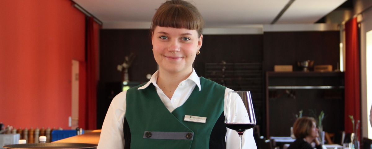 Eine Auszubildende Fachfrau für Restaurant- und Veranstaltungsgastronomie von Schloss Wackerbarth steht im stilvoll eingerichteten Restaurantbereich. In traditioneller, grüner Berufsbekleidung hält sie ein gefülltes Weinglas in der Hand und verkörpert mit ihrem freundlichen Auftreten Professionalität und Gastfreundschaft.