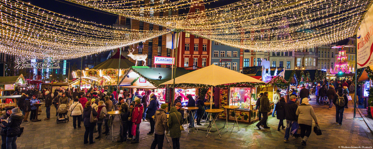 Zu sehen ist der hell erleuchtete Weihnachtsmarkt Rostock mit ganz vielen kleinen Ständen. Auch Schloss Wackerbarth ist vertreten. 